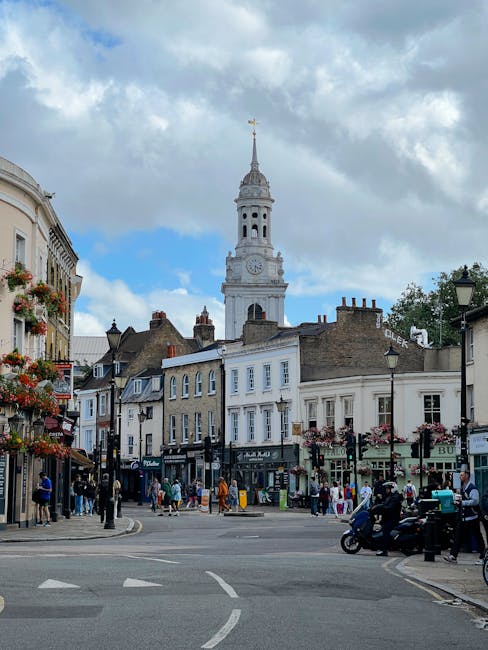 A street scene in Roman Road Market, Bow, showing a row of shops and pedestrians on the pavement. In the background, there is a historic white clock tower with a spire, set against a partly cloudy sky