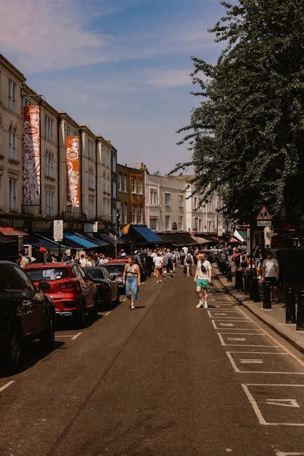A street scene in Roman Road Market, Bow, showing a row of shops and pedestrians on the pavement. In the background, there is a historic white clock tower with a spire, set against a partly cloudy sky