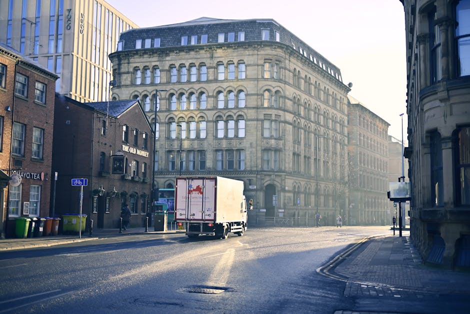 View of a busy urban street near Bow Road Station during daylight, with several vehicles including a white car, black van, and various buses moving through an intersection. The scene features a histor